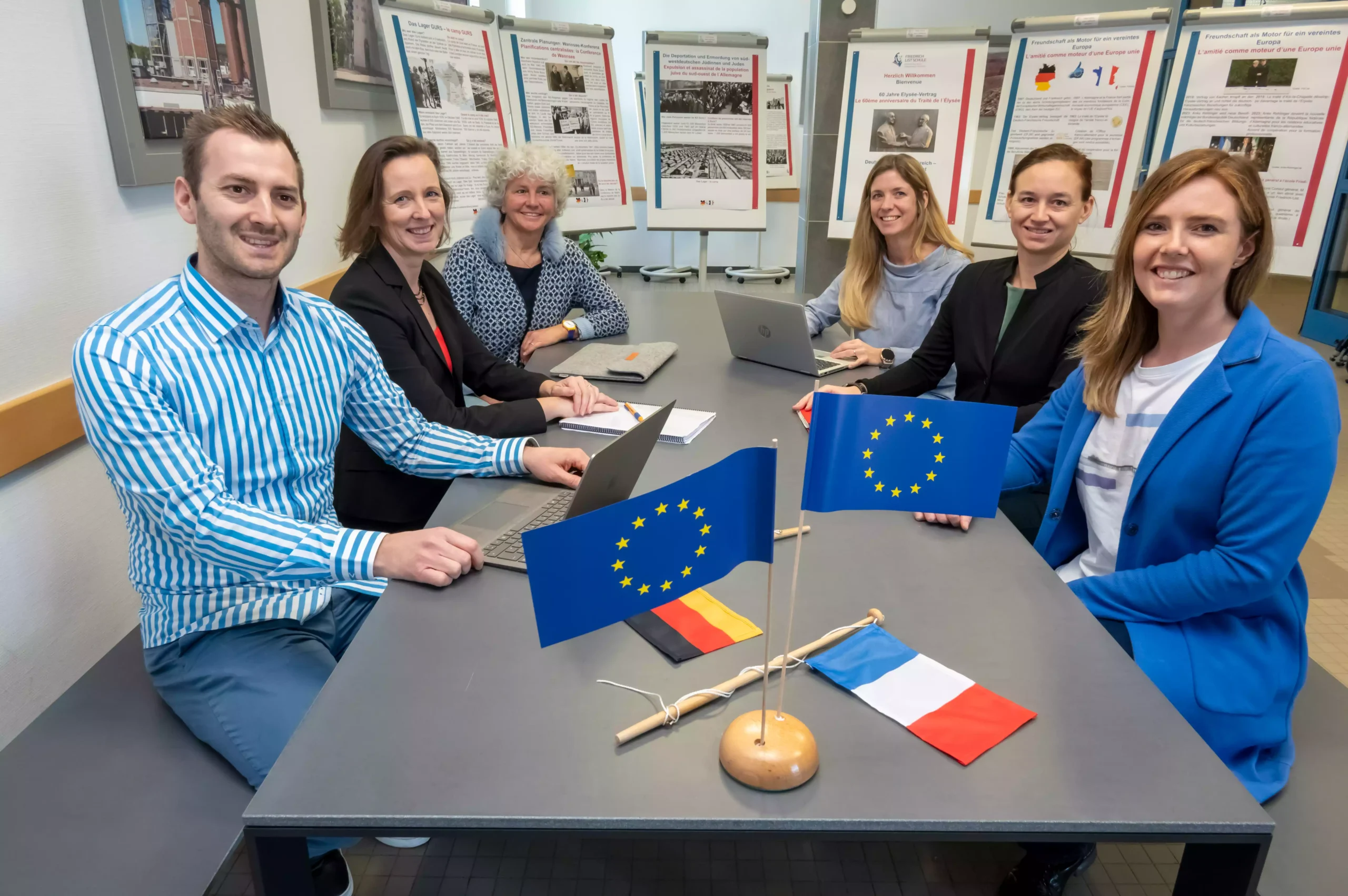 Europa Lehrkräfte-Team sitz zusammen am Tisch. Vor ihnen weht die Europa, Deutschland und Frankreich Fahne.